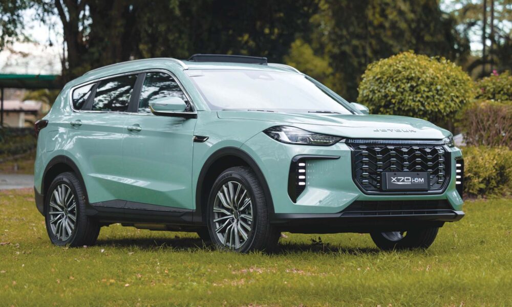 A mint-green Jetour X70 i-DM SUV parked on a grassy area, highlighting its bold front grille and sleek LED lighting design.