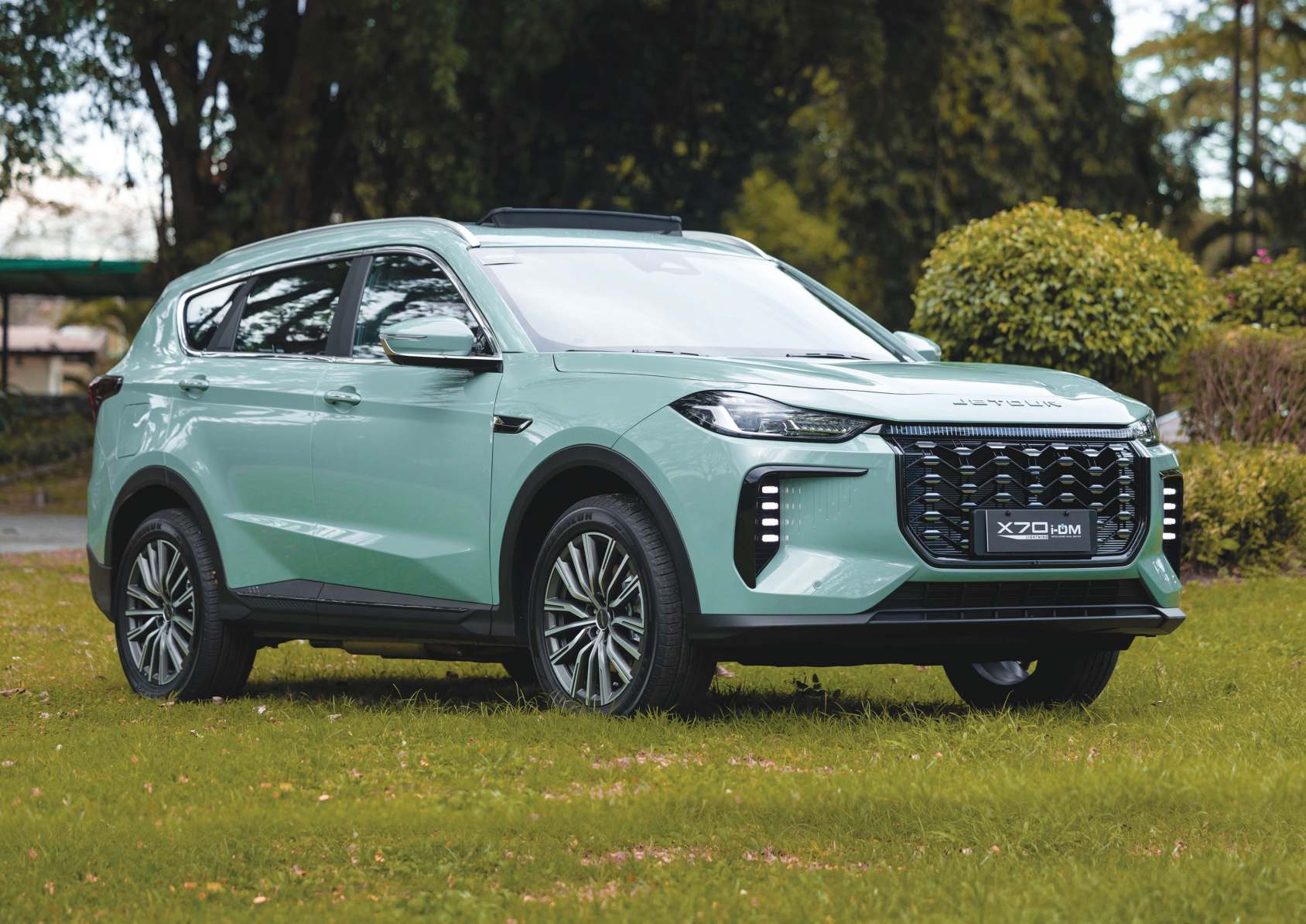 A mint-green Jetour X70 i-DM SUV parked on a grassy area, highlighting its bold front grille and sleek LED lighting design.