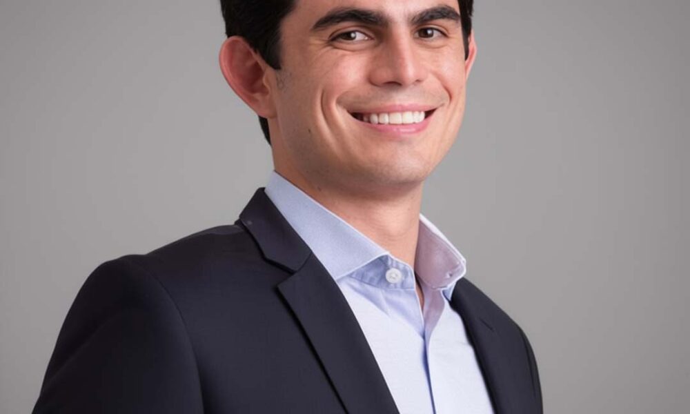 A man in a dark suit and light blue shirt smiles confidently against a neutral background.