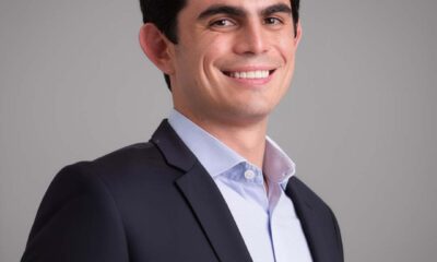 A man in a dark suit and light blue shirt smiles confidently against a neutral background.